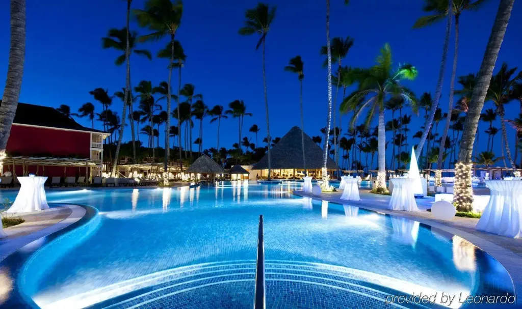 Une piscine illuminée au crépuscule, bordée de palmiers et de structures décoratives.