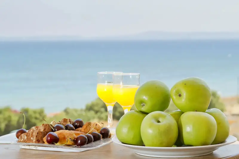 Une table est dressée avec des fruits et des viennoiseries devant une vue sur la mer.
