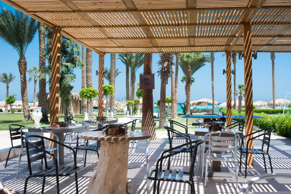 Terrasse ombragée avec tables et chaises, palmiers et vue sur la mer en arrière-plan.