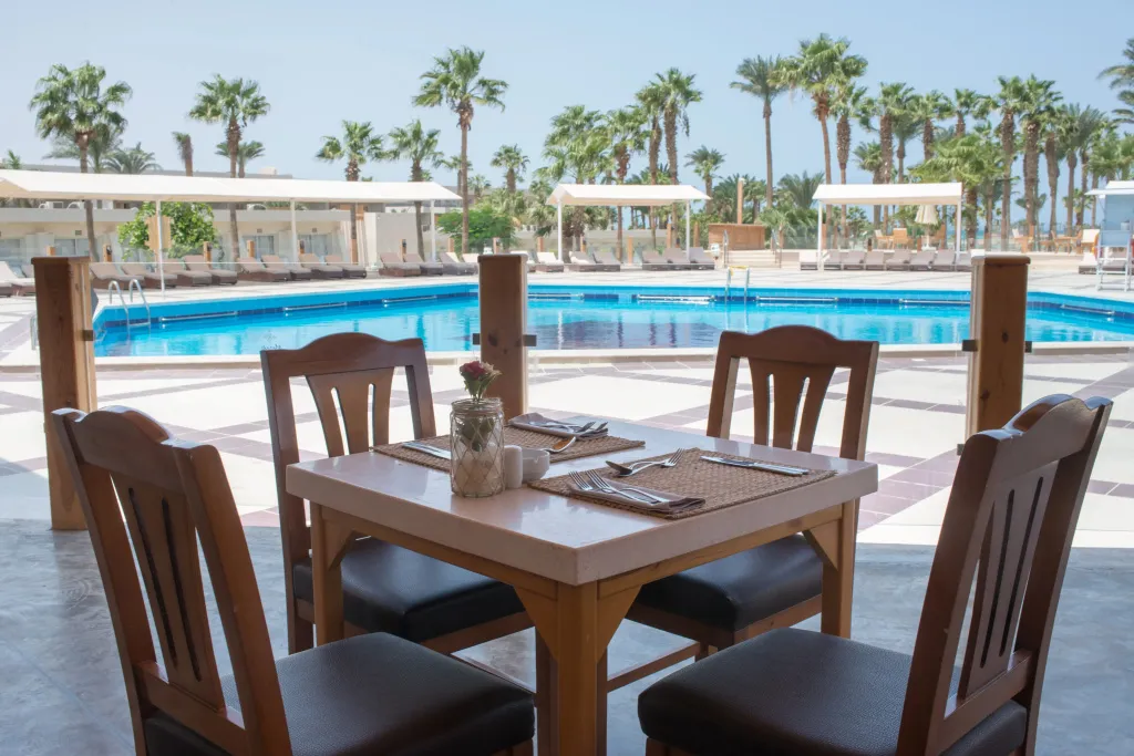 Une table est dressée devant une piscine ensoleillée, entourée de palmiers.