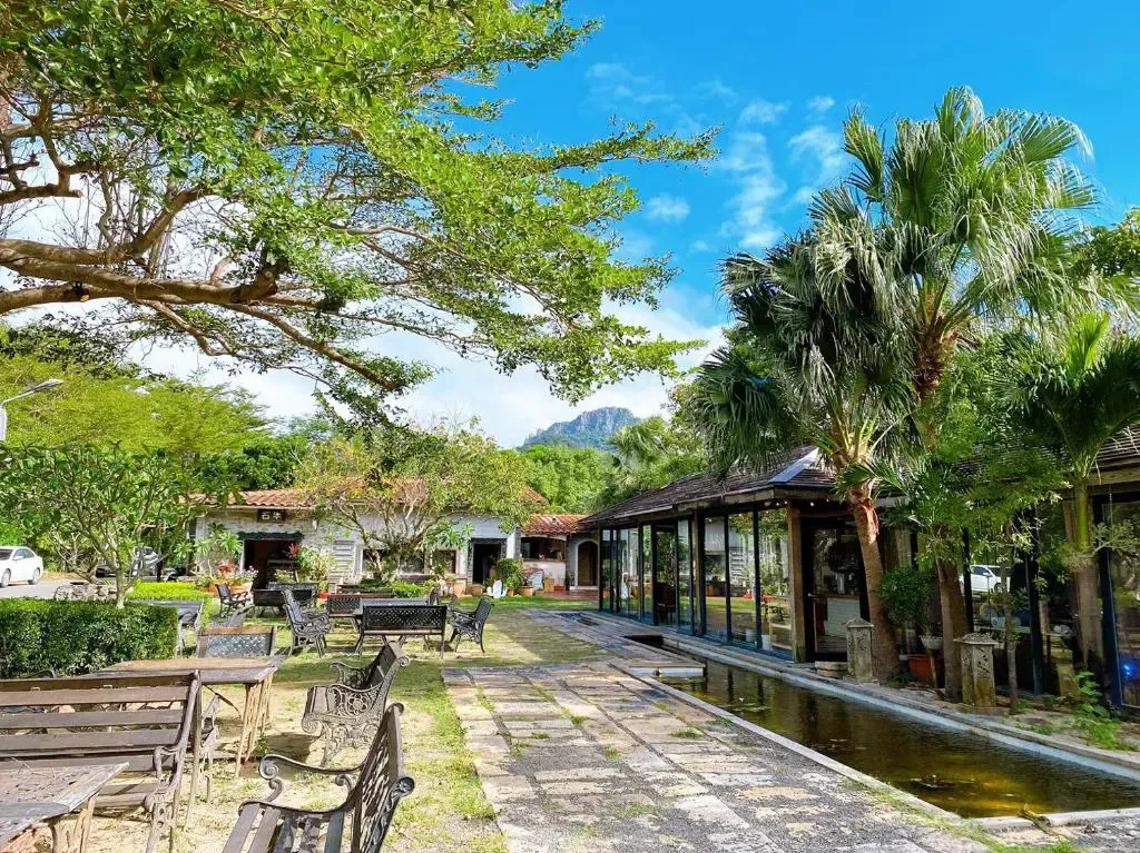Allée bordée de tables et de chaises menant à un bâtiment avec une piscine et une végétation luxuriante.