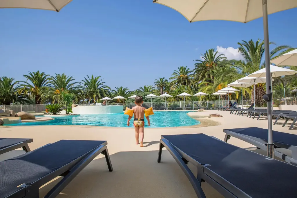 Un enfant sort d'une piscine ensoleillée, des transats invitent à la détente.