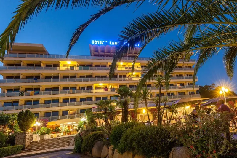 Bâtiment illuminé avec de nombreuses fenêtres et balcons, entouré de végétation tropicale au crépuscule.