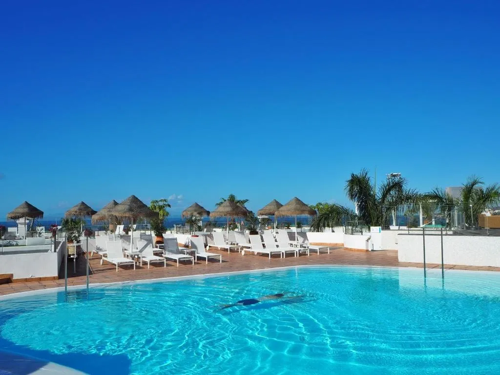 Une piscine turquoise bordée de transats sous des parasols de paille, avec une vue sur la mer.