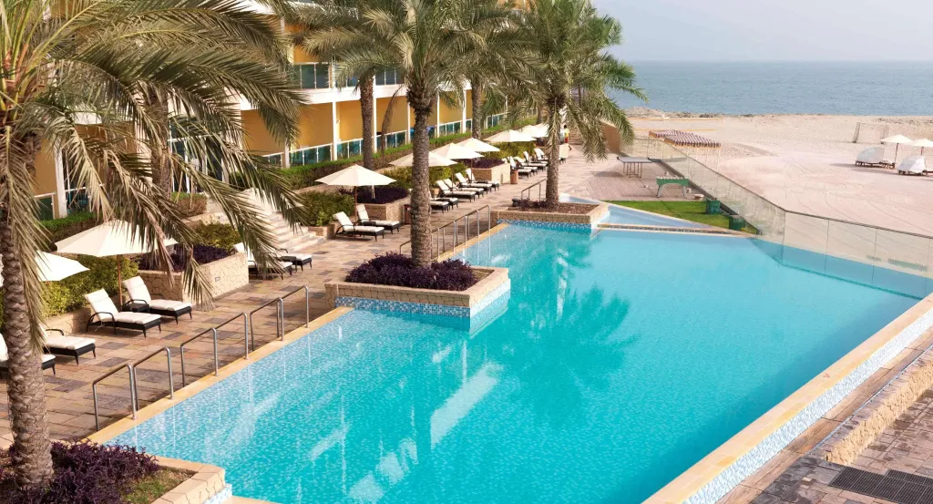 Une piscine turquoise bordée de transats et de palmiers, avec une plage en arrière-plan.