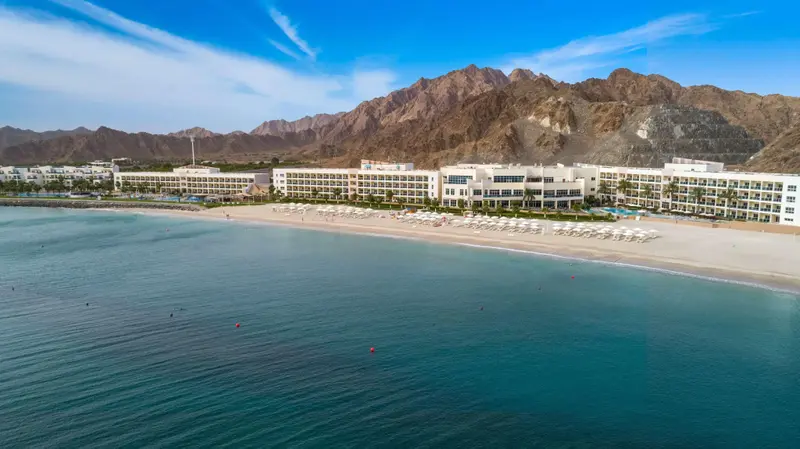 Vue aérienne d'un complexe hôtelier blanc bordant une plage de sable fin et une mer turquoise, avec des montagnes en arrière-plan.