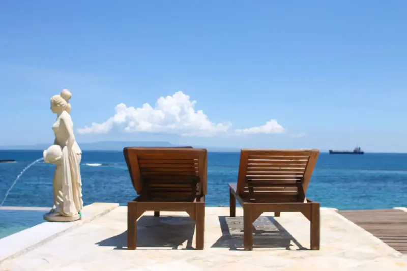 Deux chaises longues en bois se trouvent sur une terrasse ensoleillée face à la mer.
