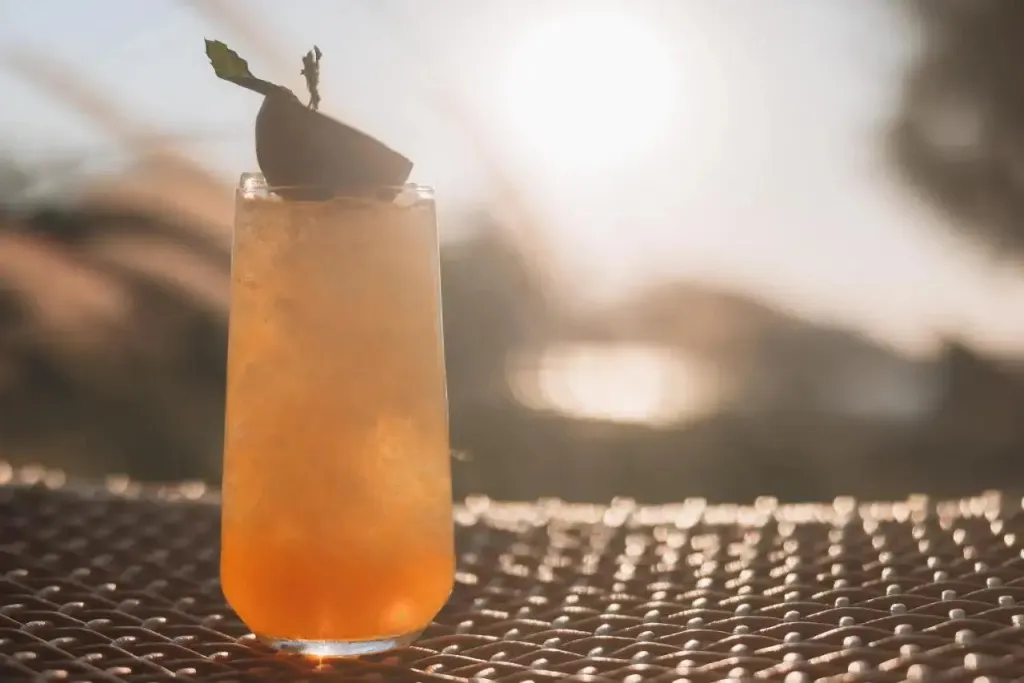 Un cocktail orangé est servi sur une table en osier avec une vue floue de la mer en arrière-plan.