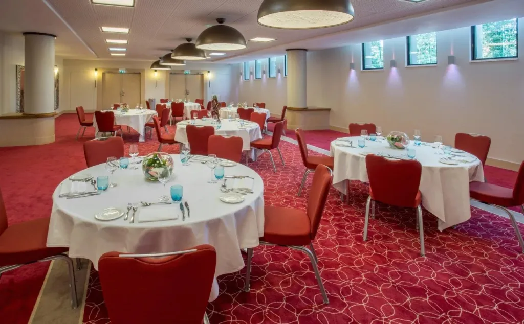 Une salle lumineuse est aménagée avec des tables rondes nappées et des chaises rouges.