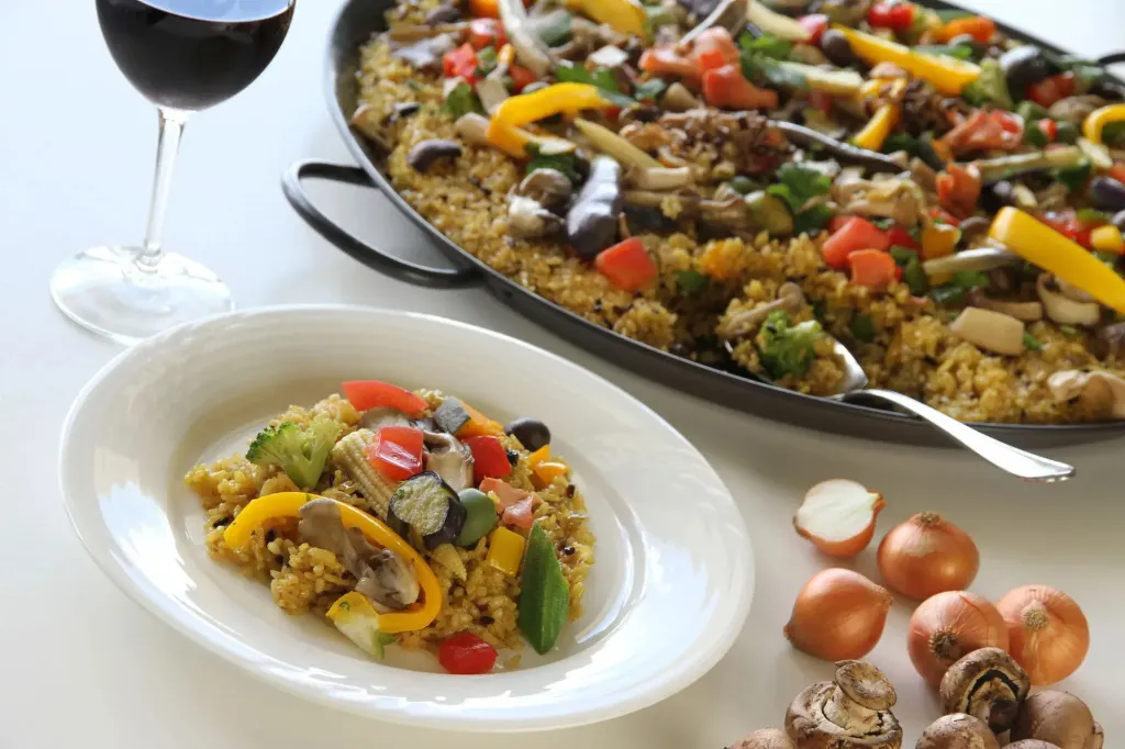 Assiette de plat préparé avec des légumes et une poêle de paella en arrière-plan.