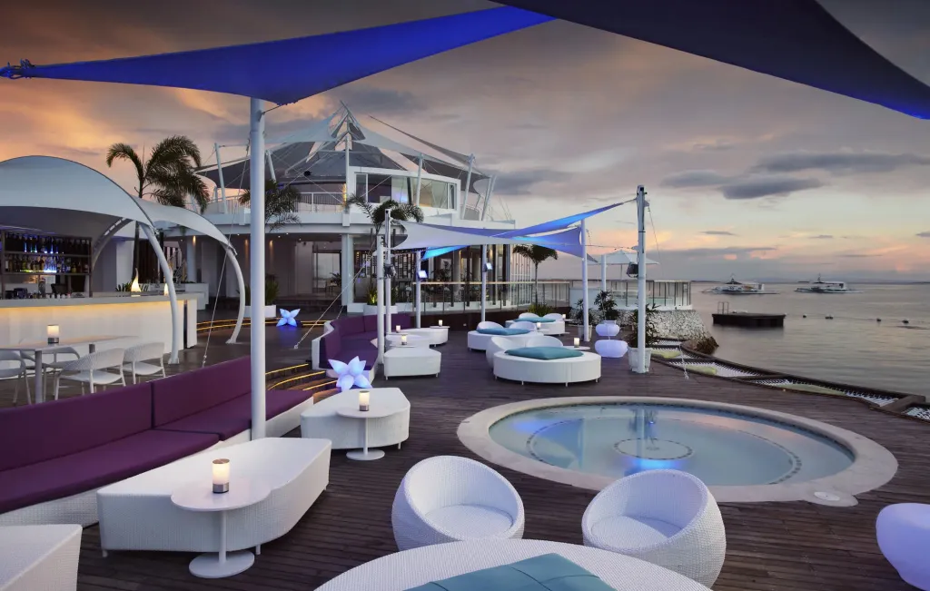 Terrasse lumineuse avec mobilier blanc et bleu, donnant sur la mer au coucher du soleil.