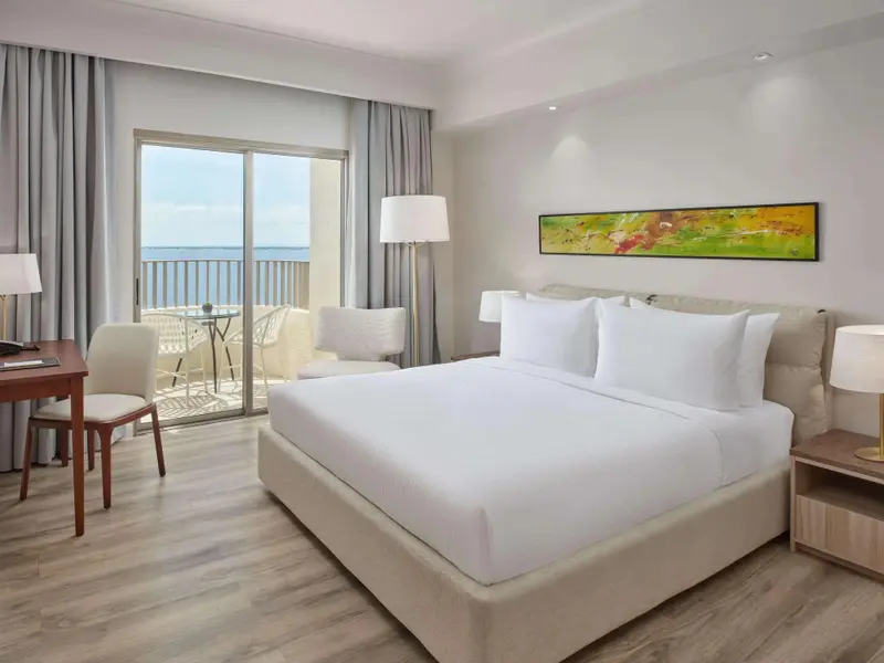 Chambre lumineuse avec un grand lit et une vue sur la mer depuis un balcon.
