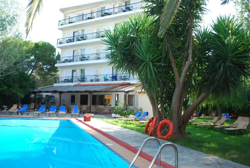 Une piscine turquoise bordée de transats, devant un bâtiment blanc à balcons.