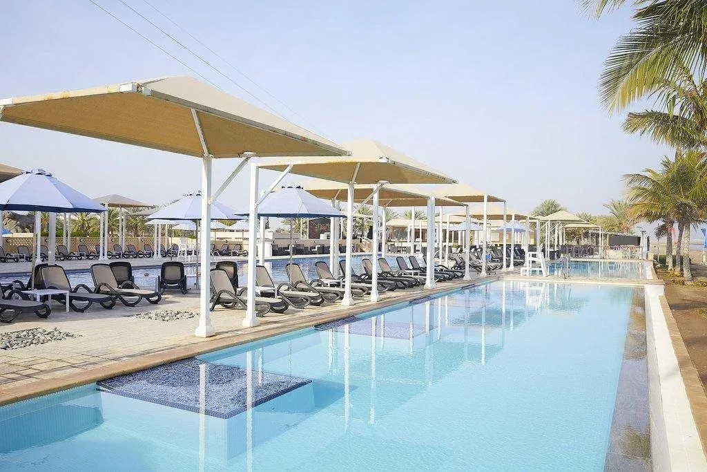 Une piscine rectangulaire bordée de transats sous des parasols beiges et bleus.