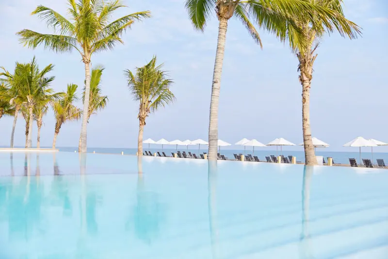 Une piscine à débordement reflète des palmiers et des transats sur une plage.