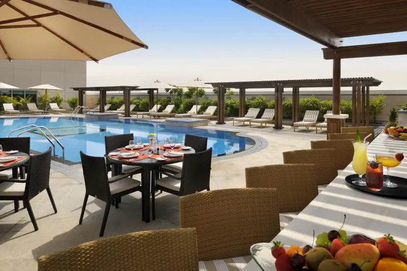 Tables dressées au bord d'une piscine ensoleillée, avec des transats et des parasols à l'arrière-plan.