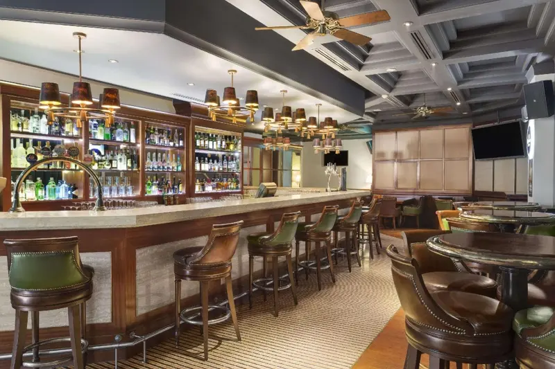 Un long comptoir de bar en bois est aligné de tabourets, avec des bouteilles exposées en arrière-plan.