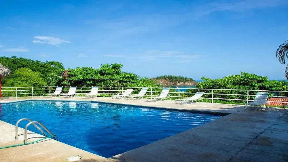 Une piscine bleue bordée de chaises longues offre une vue sur la mer et la végétation.