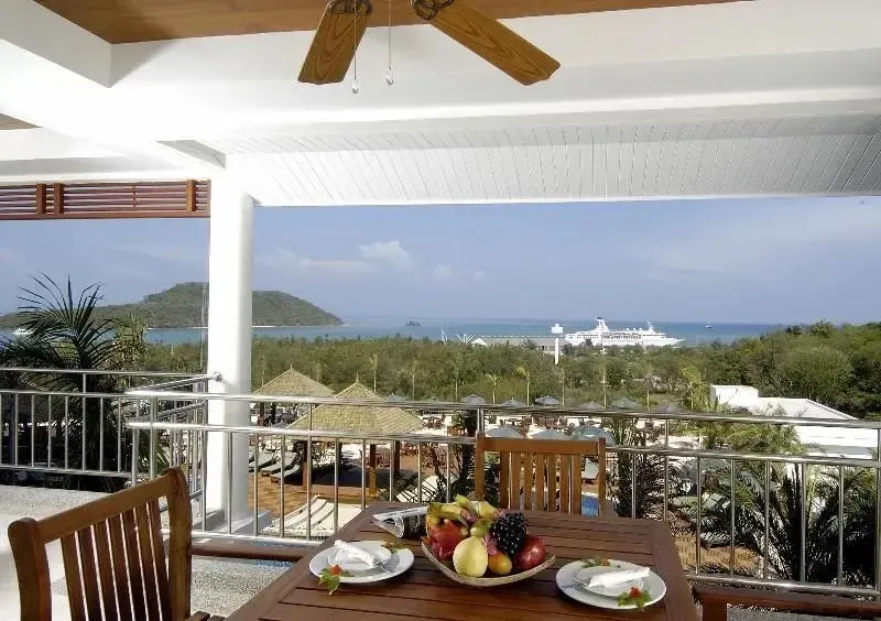 Une table de repas est mise en scène sur une terrasse ombragée avec vue sur la mer et la végétation.