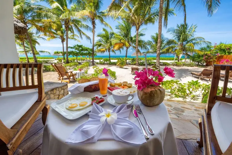 Une table est dressée avec un petit-déjeuner face à une plage de sable blanc bordée de palmiers.