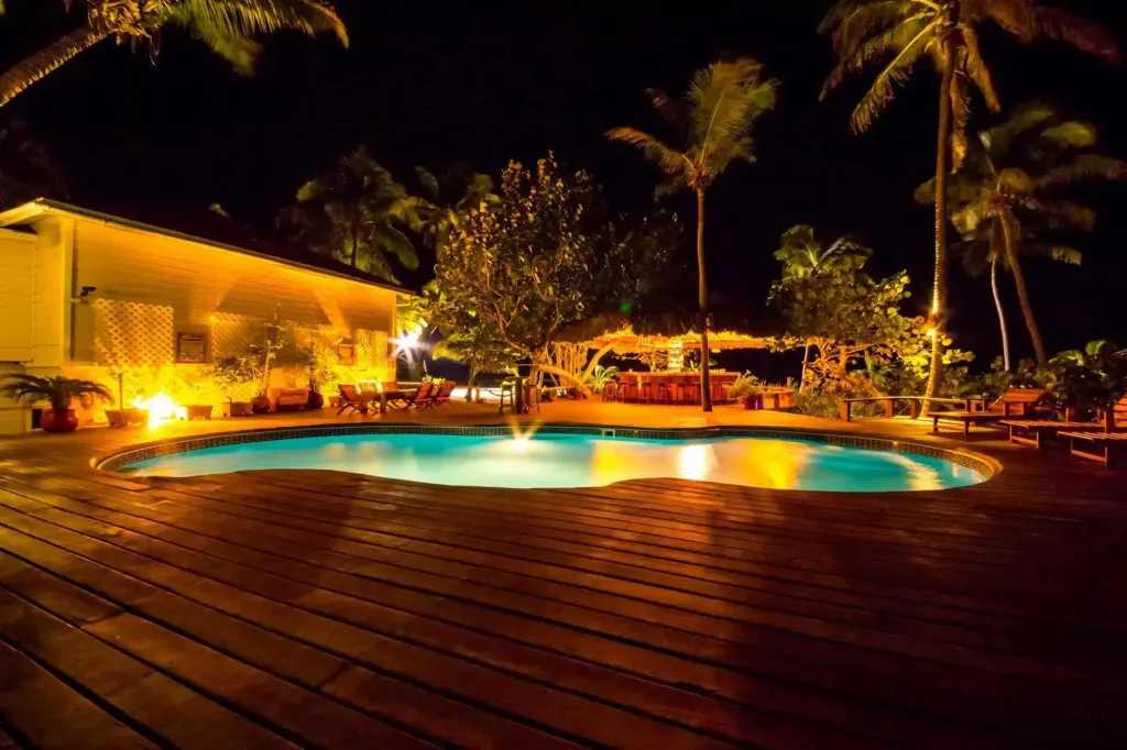 Une piscine illuminée au crépuscule, entourée d'une terrasse en bois et de végétation tropicale.