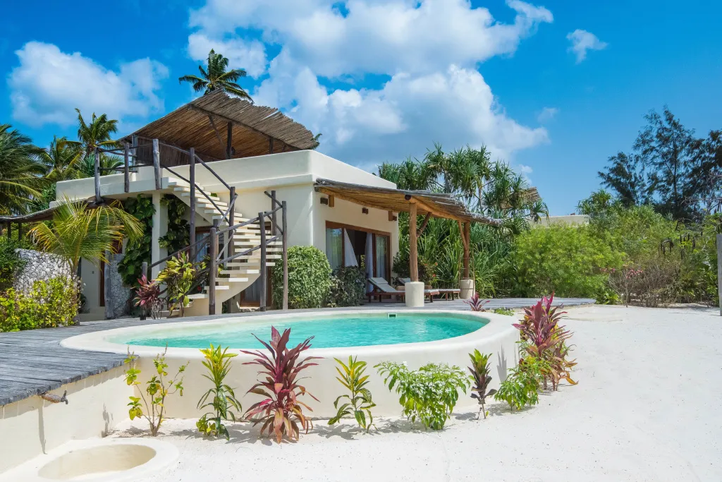 Une villa blanche avec piscine privée et terrasse ombragée se trouve sur une plage de sable blanc.