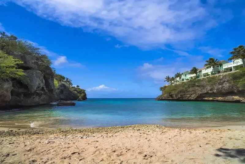 Une plage de sable fin bordée de falaises verdoyantes et d'une mer turquoise sous un ciel bleu nuageux.
