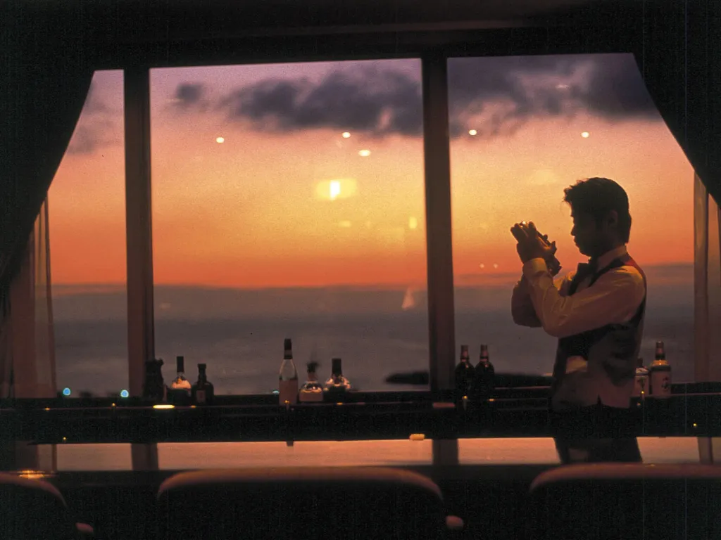 Un barman prépare un cocktail devant une vue sur la mer au coucher du soleil.