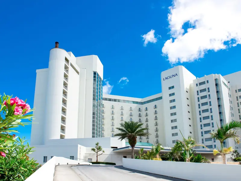 Vue d'un bâtiment blanc moderne avec des palmiers au premier plan sous un ciel bleu.