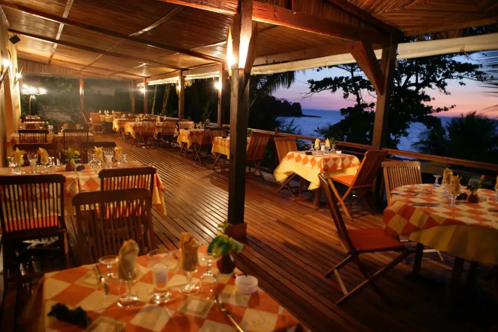Terrasse de restaurant avec tables dressées face à la mer au coucher du soleil.