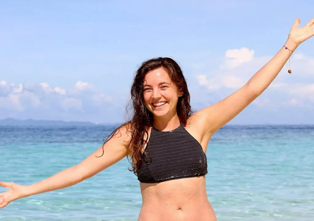 Une femme souriante se tient dans l'eau turquoise, les bras ouverts.