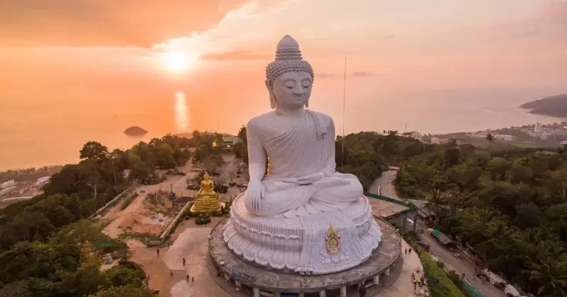 Une grande statue de Bouddha blanc domine un paysage côtier au coucher du soleil.
