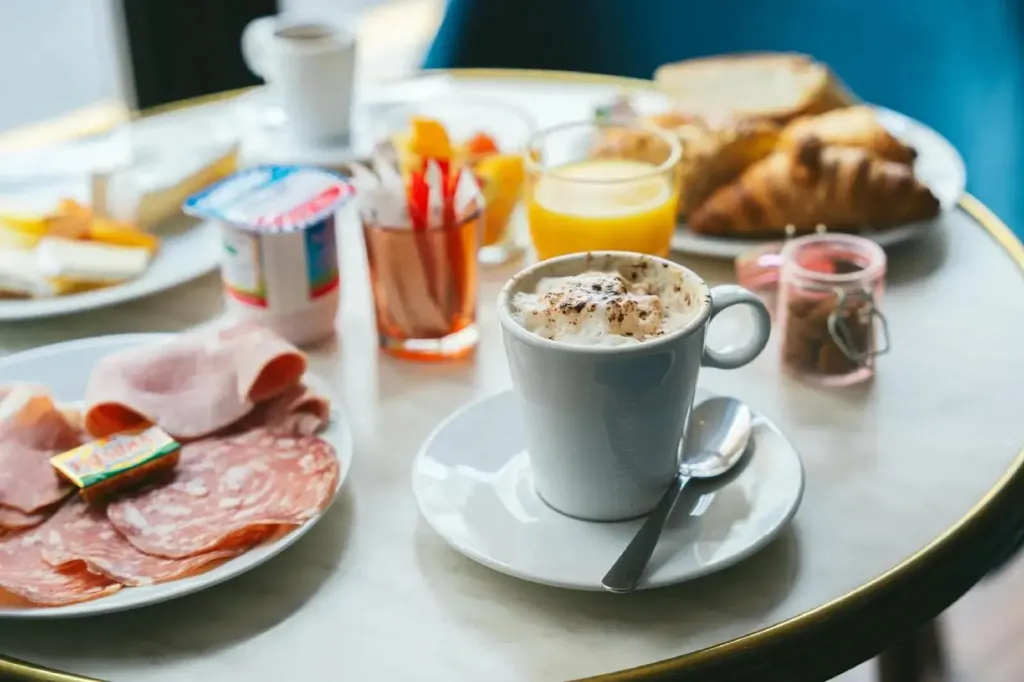 Une table ronde est dressée avec un petit-déjeuner composé de charcuterie, de viennoiseries et de boissons.