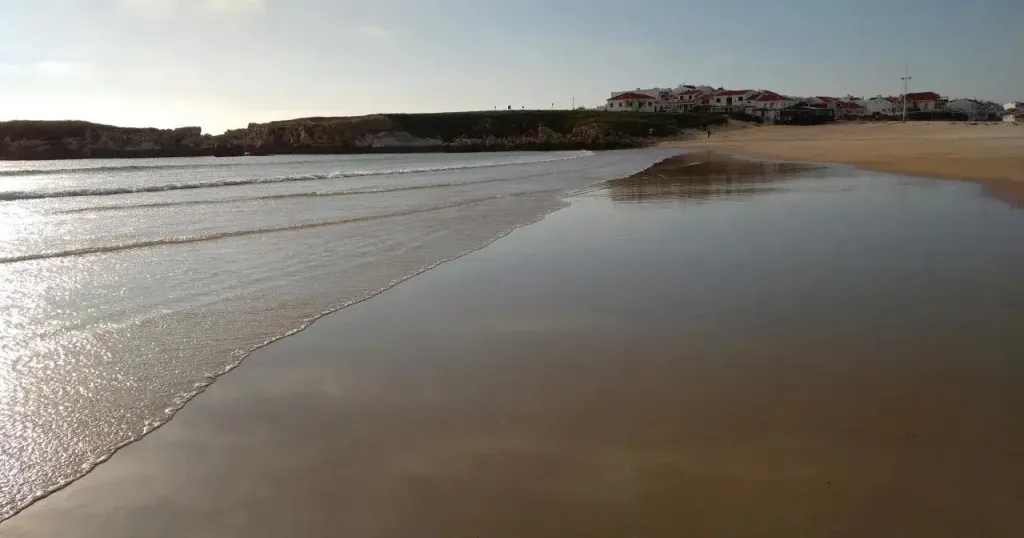 Vague se brisant sur une plage de sable fin avec des bâtiments en arrière-plan.