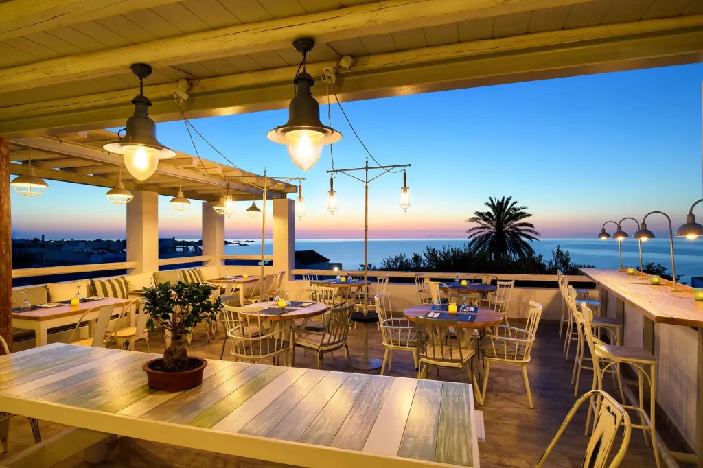 Terrasse ombragée avec tables et chaises donnant sur la mer au coucher du soleil.