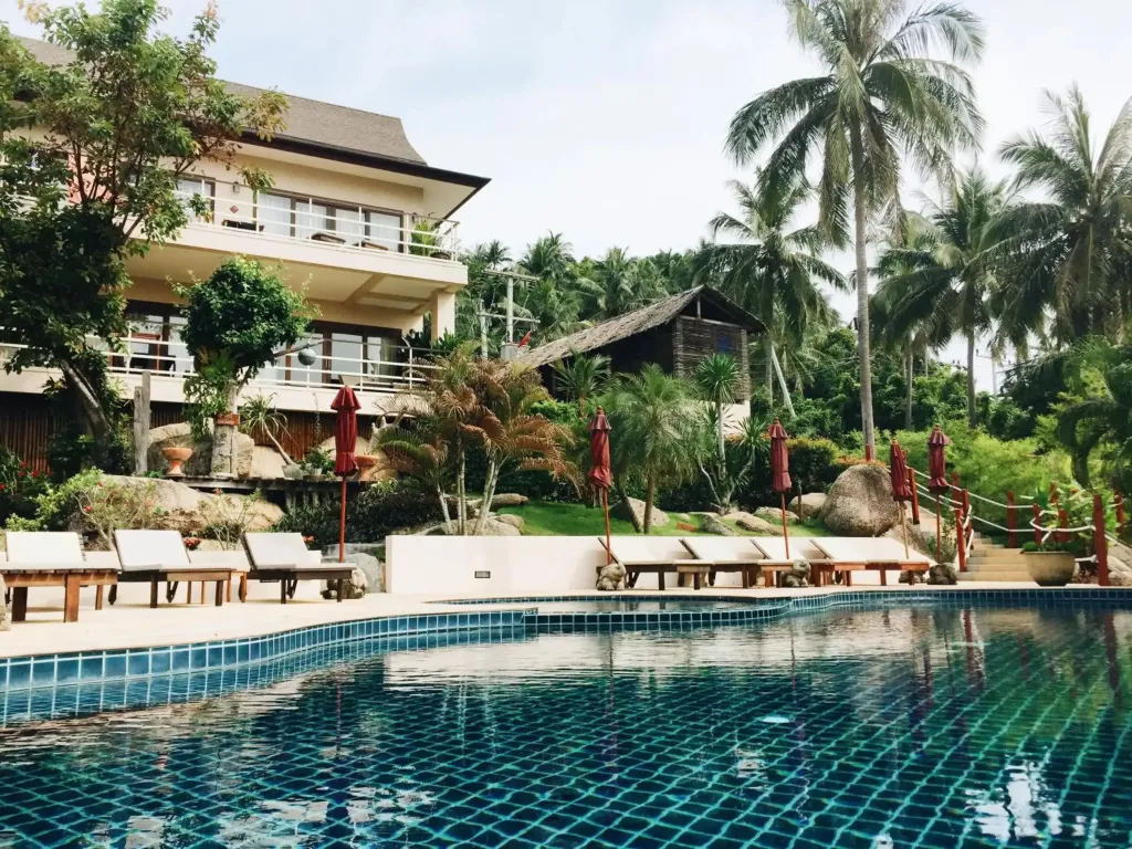 Une piscine turquoise bordée de transats, avec des bâtiments et une végétation luxuriante en arrière-plan.