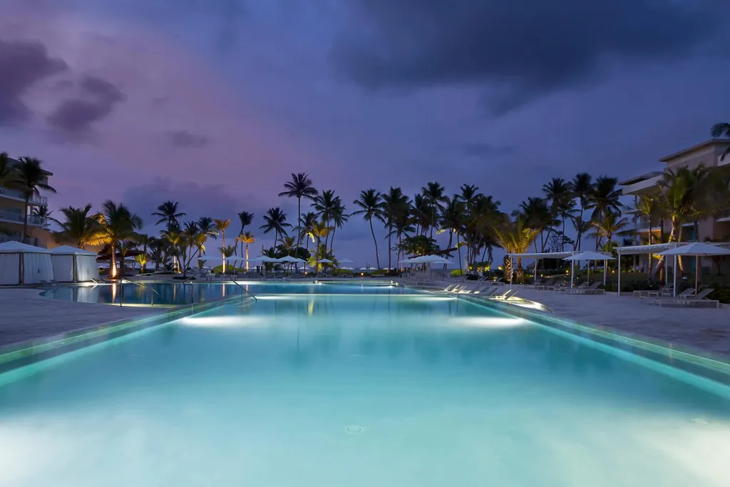 Une piscine illuminée au crépuscule, bordée de palmiers et de transats.
