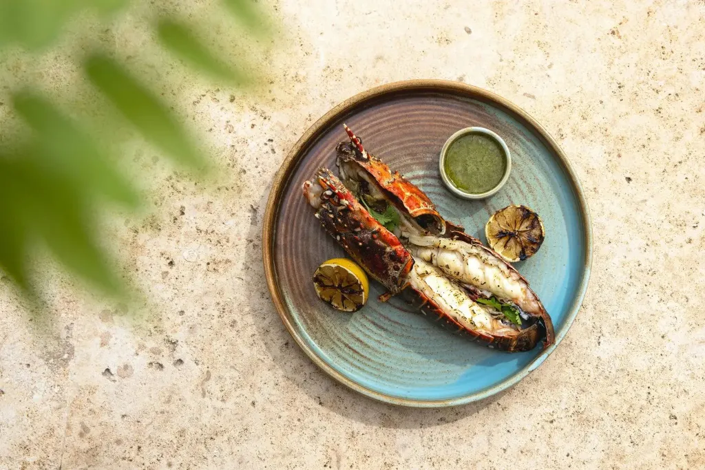 Un homard grillé est servi avec des quartiers de citron et une sauce verte sur une assiette colorée.