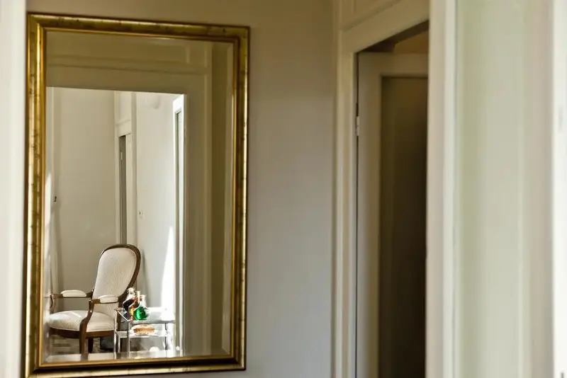 Un grand miroir doré reflète une chaise et une table d'appoint dans une pièce lumineuse.