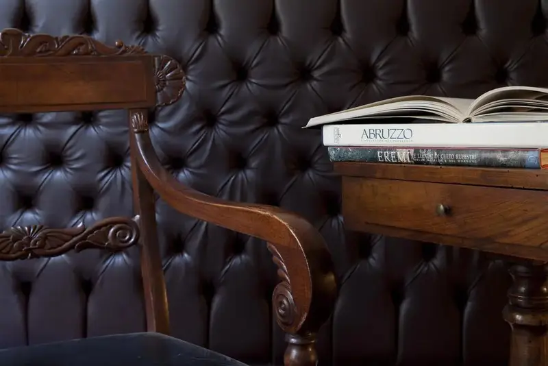 Une chaise en bois est placée devant une tête de lit capitonnée en cuir brun, avec des livres posés sur une petite table.