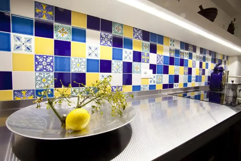 Un plan de travail en inox est devant un mur recouvert de carreaux de céramique aux motifs colorés.