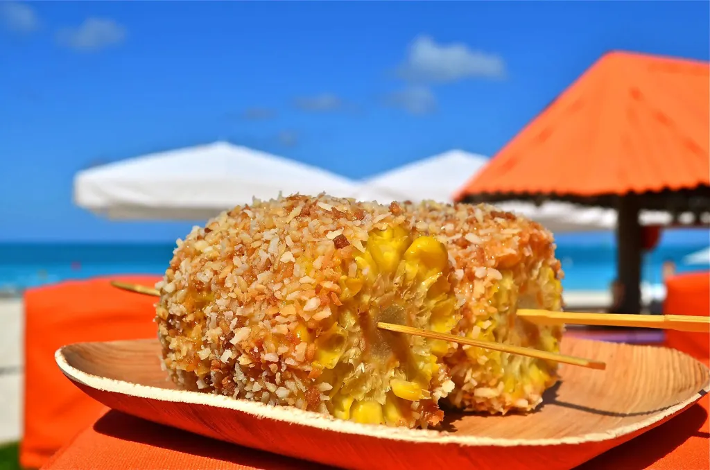 Un épi de maïs grillé, recouvert de fromage et de noix, est servi sur une assiette en bois.