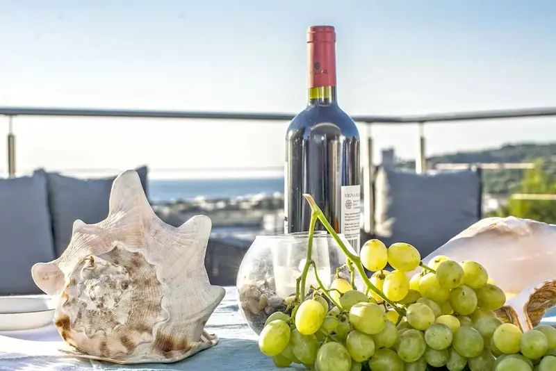 Une bouteille de vin, des raisins et des coquillages sont disposés sur une table avec vue sur la mer.