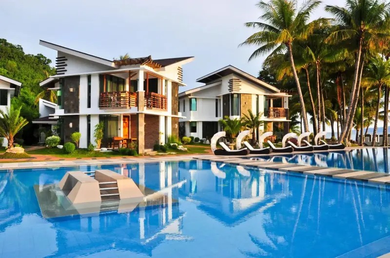 Bâtiments blancs modernes bordent une piscine turquoise et une plage tropicale.