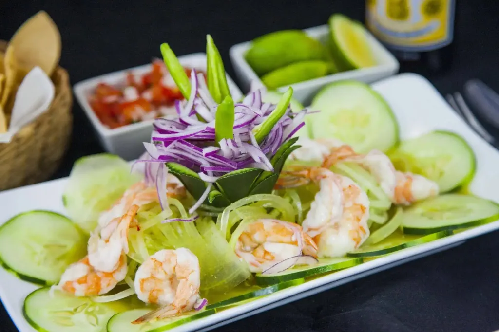 Assiette de crevettes marinées avec concombre et oignons, accompagnée de quartiers de citron vert et d'une sauce.