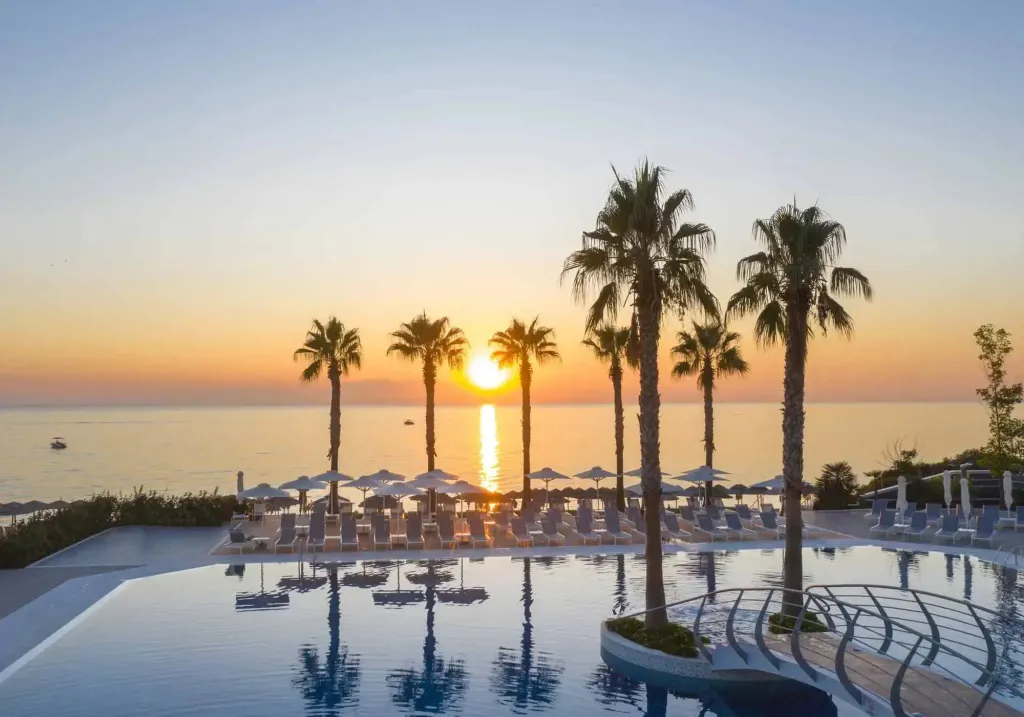 Une piscine bordée de transats et de palmiers au coucher du soleil.