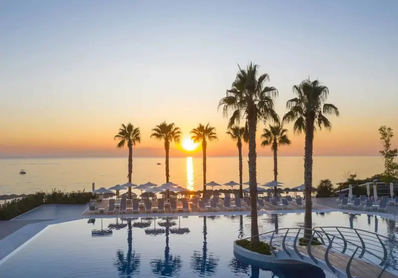 Une piscine bordée de transats et de palmiers au coucher du soleil.