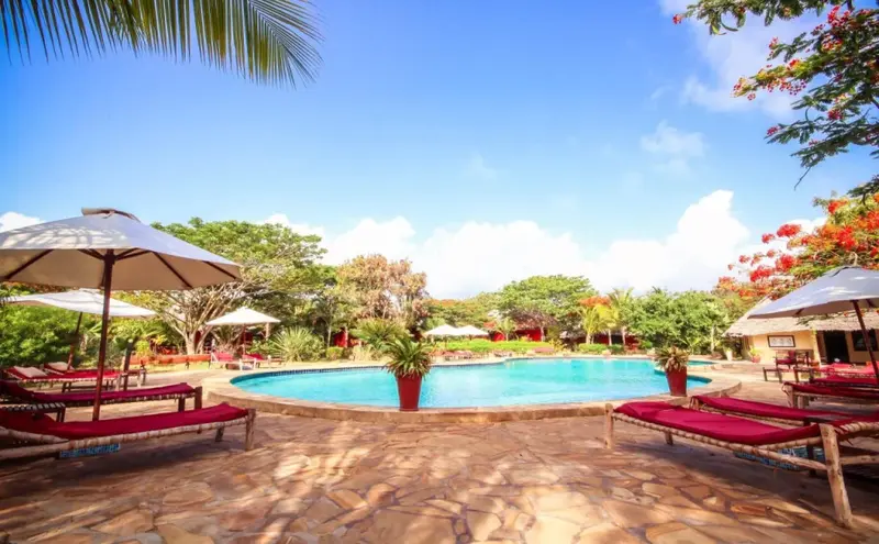 Transats rouges et parasols blancs bordent une piscine turquoise dans un cadre tropical.
