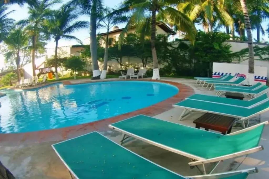Transats verts disposés autour d'une piscine turquoise bordée de végétation tropicale.