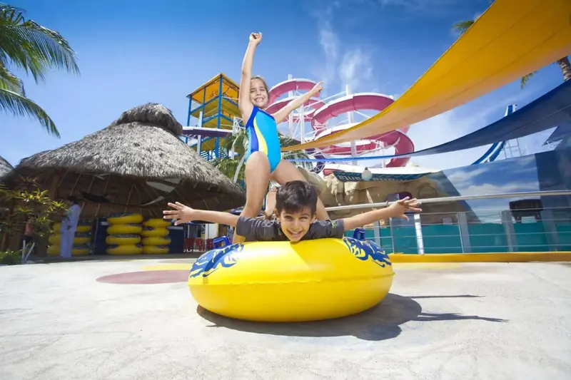 Des enfants s'amusent sur une bouée jaune devant un parc aquatique coloré.